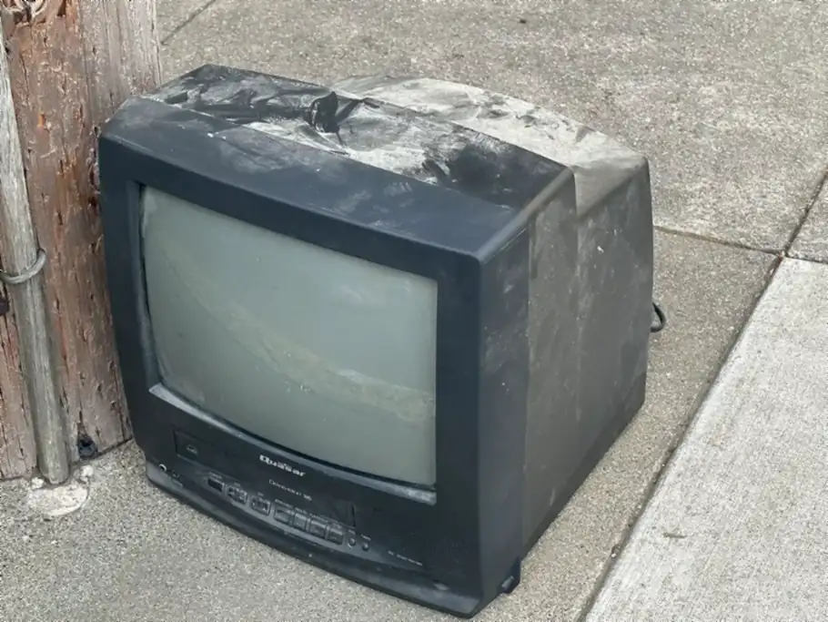 A small, dusty black tube TV sitting on a sidewalk next to a wooden utility pole