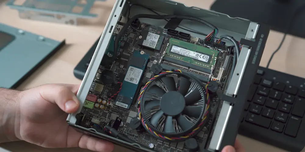 Hands holding an opened mini PC, showing a laptop-style memory stick, an M.2 SSD, and a round cooling fan