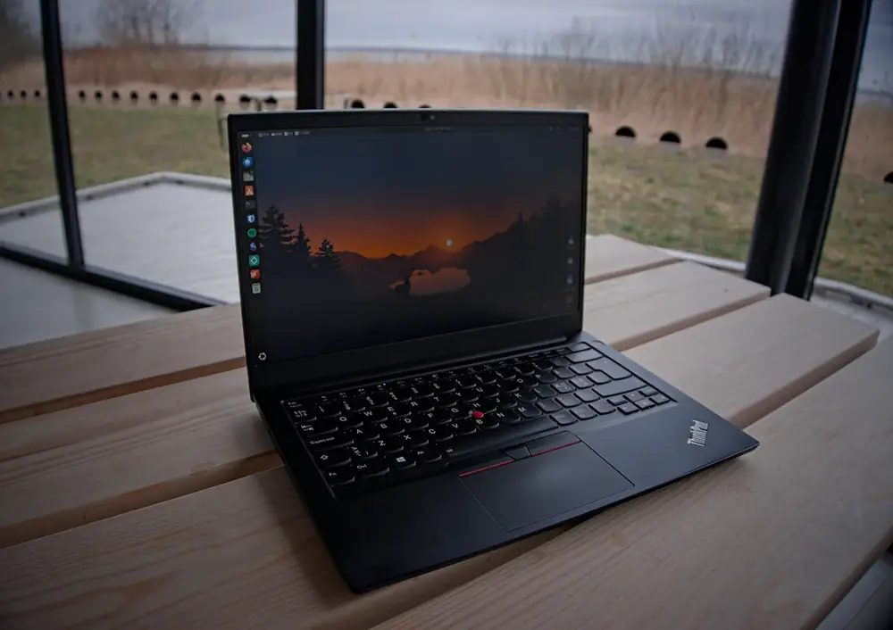 Black ThinkPad laptop open on a wooden picnic table, showing a Linux desktop with a sunset wallpaper